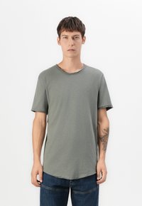 Olivenfarbener Kurzarm-T-Shirt mit lockerer Passform, Rundhalsausschnitt und rohen Kanten. Trägt dazu eine dunkelblaue Jeans, die ein Tattoo am Arm zur Schau stellt.