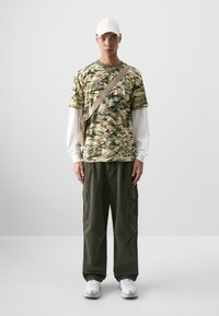 Camo-patterned t-shirt in beige with green accents, layered over a white long-sleeve shirt. Olive cargo pants and white sneakers complete the look.