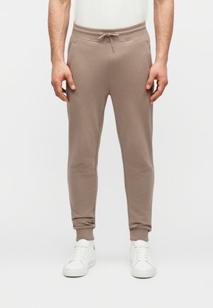 Bărbat purtând pantaloni jogger bej cu talie ajustabilă și șiret și adidași albi, stând pe un fundal simplu și deschis la culoare.