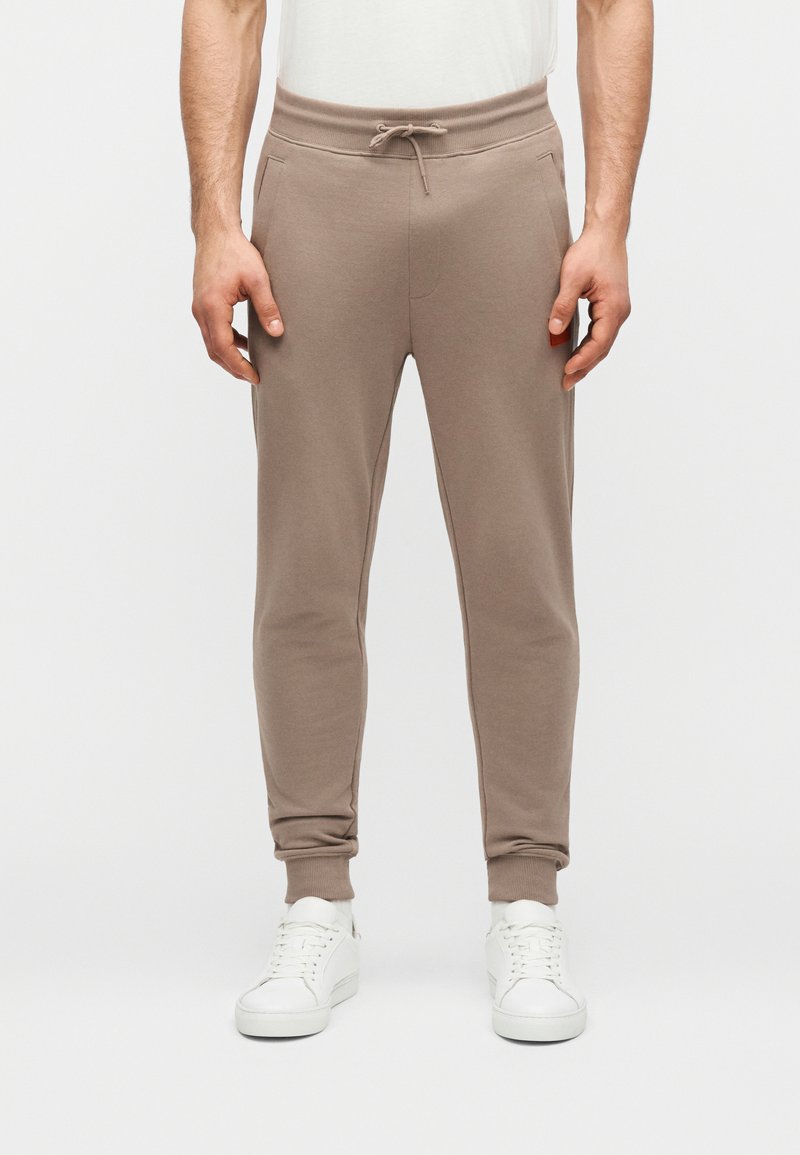 Άνδρας που φοράει μπεζ παντελόνια jogging με κορδόνι στη μέση και λευκά αθλητικά παπούτσια, στεκόμενος μπροστά σε ένα απλό φωτεινό φόντο.