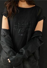 Svart kortärmad skjorta med avslappnad passform, med präglad text och rund halsringning, lager på en svart läderjacka.