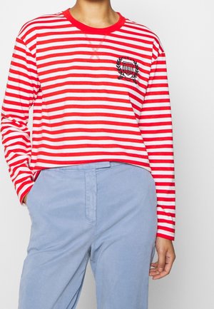 Personne portant une chemise à manches longues rayée rouge et blanche avec un petit emblème noir et un pantalon bleu clair, une main dans la poche.