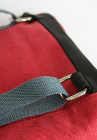 Rote Leinentasche mit glattem schwarzen Lederbesatz. Verfügt über einen grauen gewebten Riemen mit metallischen Beschlägen. Gestickte Akzente sichtbar.