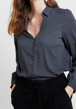 Blusa grigia scura con colletto, chiusura frontale a bottoni con bottoni rivestiti in tessuto, abbinata a pantaloni neri con vita elasticizzata.