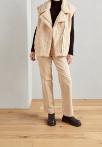 Beige Kunstpelz-Weste mit Kragen und Reißverschluss, über einem braunen Rollkragenpullover getragen, kombiniert mit hellbeigen Hosen und schwarzen klobigen Stiefeln.