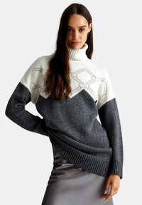 Maglione a maglia grossa con collo alto, parte superiore bianca con motivo geometrico e sezione inferiore grigio scuro. Orlo e polsini a costine.