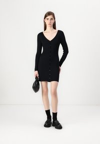Robe noire en tricot côtelé avec des manches longues et un col en V. Présente une rangée de boutons sur le devant. Associée à des chaussures noires chunky et un sac.