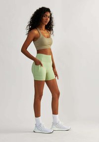 Jasnozielone krótkie legginsy z bocznymi kieszeniami, zestawione z beżowym stanikem sportowym i białymi butami sportowymi. Gładka, elastyczna tekstura materiału.