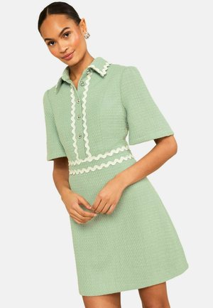 Frau trägt ein strukturiertes hellgrünes Kleid mit kurzen Ärmeln, gewelltem weißen Besatz, silbernen Knöpfen und spitzem Kragen, posiert mit gefalteten Händen.