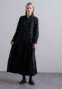 Long-sleeve, dark green plaid dress made of soft fabric, featuring a button-down front and a tiered skirt design, worn with black shoes.