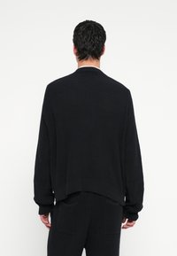 Črni pleteni pulover z udobnim krojem, rebrasto teksturo, padajočimi rameni in okroglim izrezom. Oblečen s hlačami v istem slogu z žepki na zalepko.