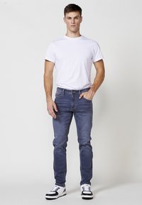 Jeans de corte skinny en lavado azul oscuro, con un diseño clásico de cinco bolsillos y detalles de costura sutiles, combinados con una camiseta blanca lisa y zapatillas negras/blancas.