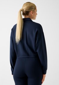 Felpa cropped blu navy con maniche lunghe e vestibilità rilassata. Tessuto liscio, senza motivi visibili e con un semplice design del colletto sul retro.