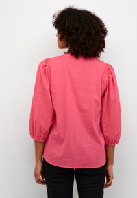 Blusa rosa con maniche a sbuffo, tessuto testurizzato con piccole gobbe, orlo arrotondato, indossata con pantaloni neri.