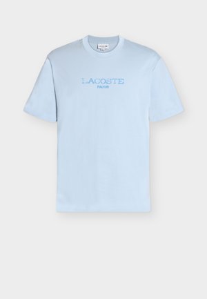 Svetlo modra majica kratkih rokavov z okroglim vratom, z izvezenim napisom "LACOSTE PARIS" v ujemajoči modri barvi na prsih.