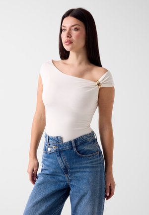 Femme aux longs cheveux foncés portant un haut blanc asymétrique avec détail anneau doré et jean bleu taille haute sur fond uni.