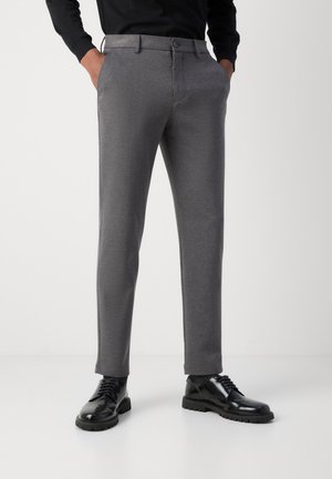 Pantaloni grigi su misura con una texture liscia, dotati di chiusura con bottone e tasche laterali. Abbinati a scarpe nere lucide.