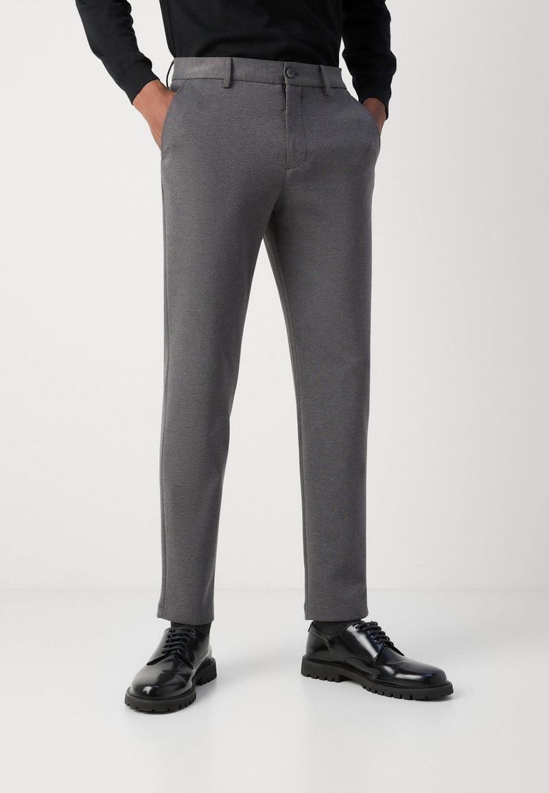 Sivo prilagojene hlače z gladko teksturo, z zapiranjem na gumb in stranskimi žepi. Kombinirane z bleščečimi črnimi čevlji.