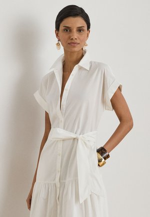 Naine, kes kannab valget nööpidega kleiti, millel on sõlmitud vöö vöökohal, suured kõrvarõngad ning randmel kuldne ja kilpkonnakujuline käevõru.