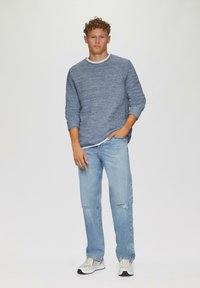 Lichtblauwe gebreide trui met een gestructureerd patroon, gecombineerd met lichtblauwe versleten spijkerbroek en witte sneakers. Casual, relaxte pasvorm.