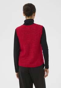 Punainen neulottu liivi, jossa on tekstuuri, pyöreä pääntie ja hihaton malli, puettuna mustan pitkähihhaisen paidan päälle.