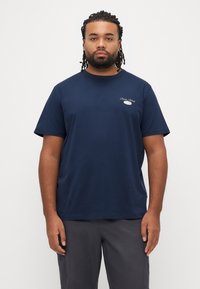 Tamsiai mėlynas medvilninis marškinėlis trumpomis rankovėmis ir apvalia iškirpte, su mažyčiu siuvinėtu logotipu ant krūtinės. Lygus audinys.