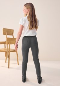 Pantalon gris coupe slim avec poches arrière, associé à un haut blanc à manches courtes, debout à côté d'une chaise en bois sur un fond neutre.