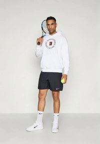 Man holding tennis racket and ball, wearing white Nike hoodie, black shorts, white socks, and white sneakers, standing against plain background.