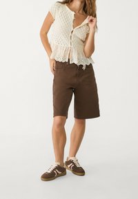 Mujer vestida con una blusa de encaje color crema abotonada, pantalones cortos marrones hasta la rodilla y zapatillas marrones con cordones y suelas blancas, de pie sobre un fondo blanco.