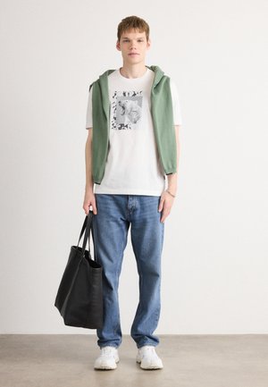 Grønn ermeløs hettegenser over en hvit grafisk t-skjorte, blå jeans og hvite sneakers, holder en svart totebag.