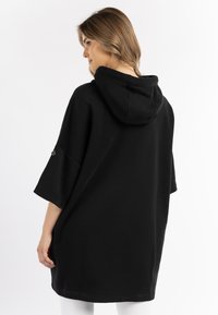 Rochie cu glugă supradimensionată, de culoare neagră, cu glugă centrală, mâneci scurte și tiv curbat. Țesătura are o textură netedă; prezintă un branding minim.