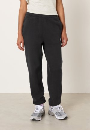 Person iført sorte sweatpants, hvide og grå sneakers og en lysefarvet kortærmet skjorte, der står på et glat gulv.