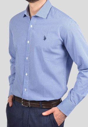 Man wearing een lichtblauw geruit overhemd met lange mouwen, ingestopt in marineblauwe broek met een donkerbruine riem, handen in de zakken.