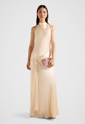 Kobieta stojąca, ubrana w długą do ziemi, bezrękawnikową suknię z różowego satynu, trzymająca małą, ozdobioną różową kopertówkę.