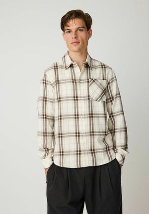 Ung mand iført en creme- og brun ternet skjorte med brystlomme og mørke løse bukser, stående med hænderne i lommerne.