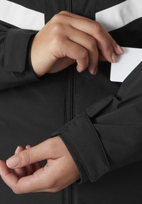 Schwarze Jacke mit weißen diagonalen Streifen, Reißverschluss und Klettverschluss an den Bündchen. Eine Hand erreicht die mit Reißverschluss versehenen Ärmeltasche und hält eine Karte.