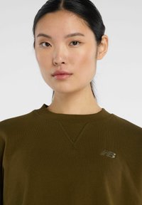 Sudadera verde oliva con escote redondo. Presenta un detalle sutil de punto en forma de V y un pequeño logo reflectante de New Balance en el pecho.