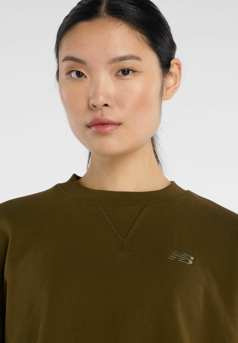 Sweat-shirt vert olive avec un col rond. Présente un discret détail en point de surjet en V et un petit logo réfléchissant New Balance sur la poitrine.