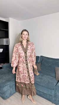 Rochie roz cu imprimeu floral, mâneci bufante, detaliu cu nasturi, modele intricate și tiv verde contrastant. Purtați cu pantofi nude cu toc, stând pe o canapea albastră.
