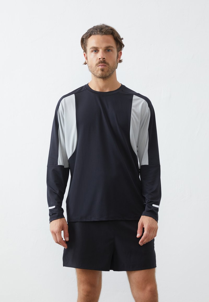 Zwarte sportieve longsleeve shirt met lichtgrijze zijpanelen, gemaakt van vochtdoorlatende stof; gecombineerd met zwarte shorts.