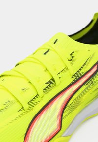 Helles gelbes Sportschuh mit strukturiertem Obermaterial, schwarzen Akzenten und reflektierenden Details. Verfügt über reguläre Schnürung und eine gepolsterte Sohle.