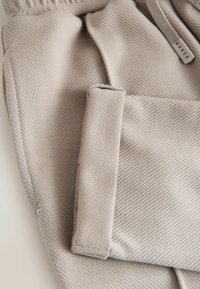 Helles beiges Gewebe mit gerippter Textur, versehen mit einer gefalteten Tasche und einem Etikett mit der Aufschrift "BAKER." Das Design ist schlicht und praktisch.