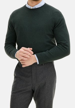 Homme portant un pull vert foncé col rond par-dessus une chemise bleu clair à col et un pantalon gris foncé, les mains jointes devant à la taille.