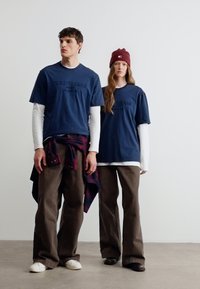 Δύο άτομα στέκονται μπροστά σε έναν λευκό τοίχο φορώντας μπλε πουκάμισα με στρώσεις, καφέ παντελόνια, αθλητικά παπούτσια και ένα κόκκινο πλεκτό σκουφάκι σε ένα από αυτά.