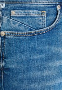 Modré denimové džíny s malou přední kapsou, kontrastním žlutým stehem a dvěma kovovými knoflíky u pasu.