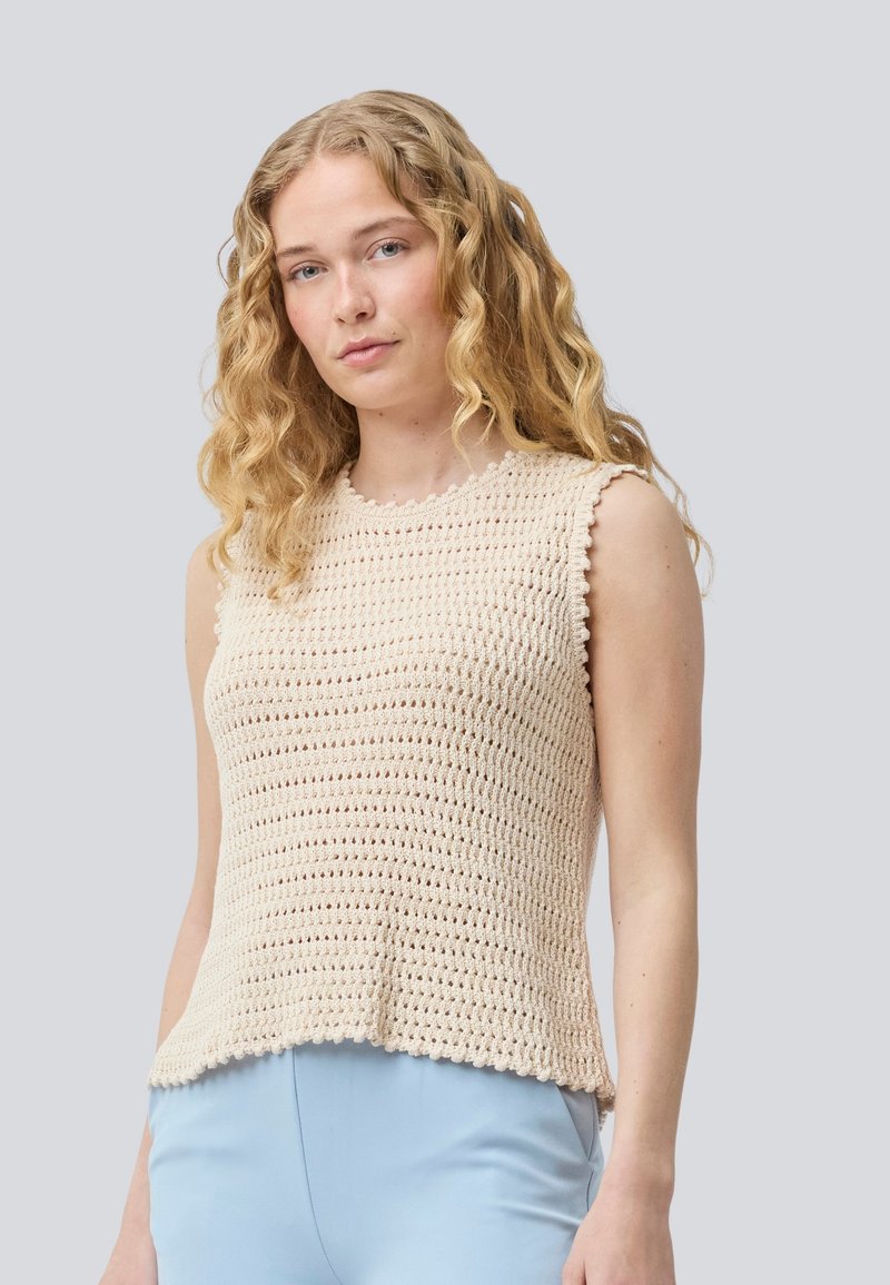 Beige kudumisest ilma varrukateta tops, millel on tekstuuriline muster ja lainelised servad, koos helesiniste pükstega, esitletud neutraalses taustas.