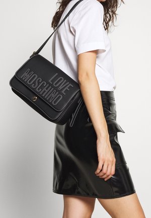 Vrouw draagt een witte blouse en een zwarte leren rok, draagt een zwarte schoudertas met de tekst "LOVE MOSCHINO" in witte stiksel.