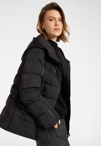 Giubbotto piumino nero con cappuccio, design trapuntato, tasche con zip e texture liscia, indossato sopra un maglione nero e pantaloni grigi.