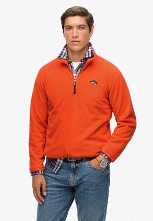 Oranger Fleece-Pullover mit einem halben Reißverschluss-Kragen, versehen mit einem kleinen Logo auf der Brust und unterbrochen von den zu sehenden Karomuster-Bündchen eines Hemdes. Kombiniert mit Jeans.