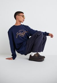 Mornarska mikica z oranžnim logotipom, kombinirana z hlačami s črtami in rjavimi čevlji z rumenim šivom. Model se sklanja, nosi sončna očala.